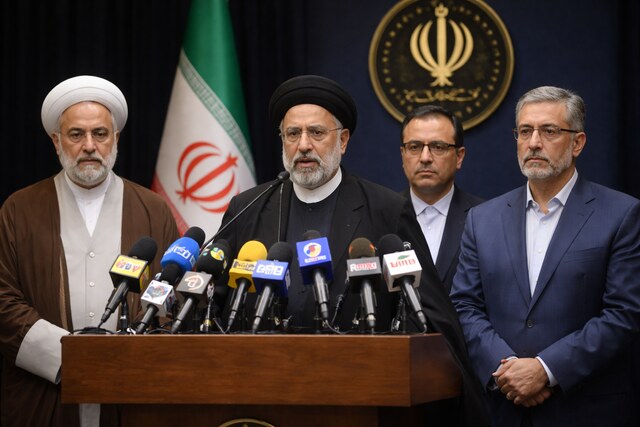 Iranian government officials seated at a press conference table during an official briefing, with microphones and national flags visible in a formal setting.